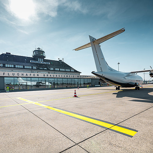 Vorfeld des Flughafens Braunschweig-Wolfsburg bei sonnigem Wetter. Rechts steht ein wei&szlig;es Turboprop-Flugzeug, im Hintergrund sind das Terminal mit der Aufschrift &bdquo;FLUGHAFEN BRAUNSCHWEIG-WOLFSBURG&ldquo; und der Kontrollturm zu sehen. Markierungen auf der Betonfl&auml;che f&uuml;hren durch das Bild, dar&uuml;ber spannt sich ein klarer blauer Himmel mit leichter Bew&ouml;lkung.